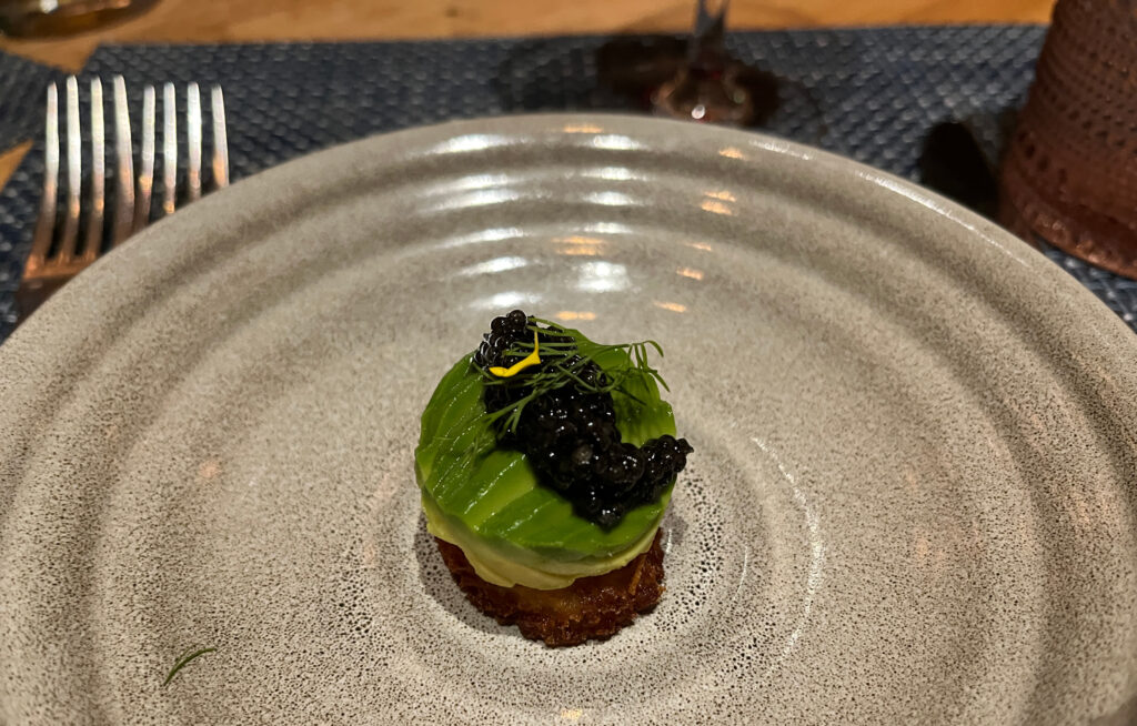 Small potato latke topped with sliced avocado and caviar, on a gray plate.