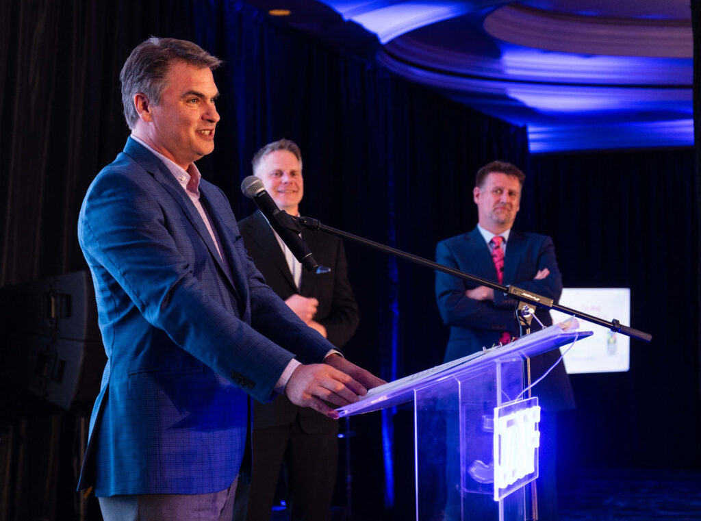 Dan Connors gives his award acceptance speech, with Josh Larson and Hans ten Cate watching in the background.