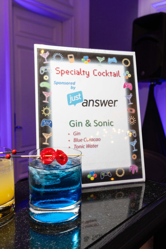 Blue drink with maraschino cherries at the top, in front of a sign that says "Specialty Cocktail sponsored by JustAnswer. Gin & Sonic: Gin, Blue Curacao, Tonic Water"