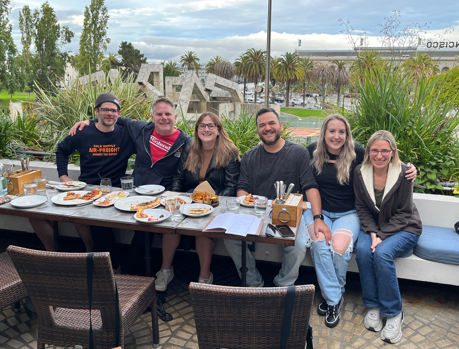 A group of people sitting at an outdoor table with food and drinks on it, with the sky and Port of San Francisco building in the background.