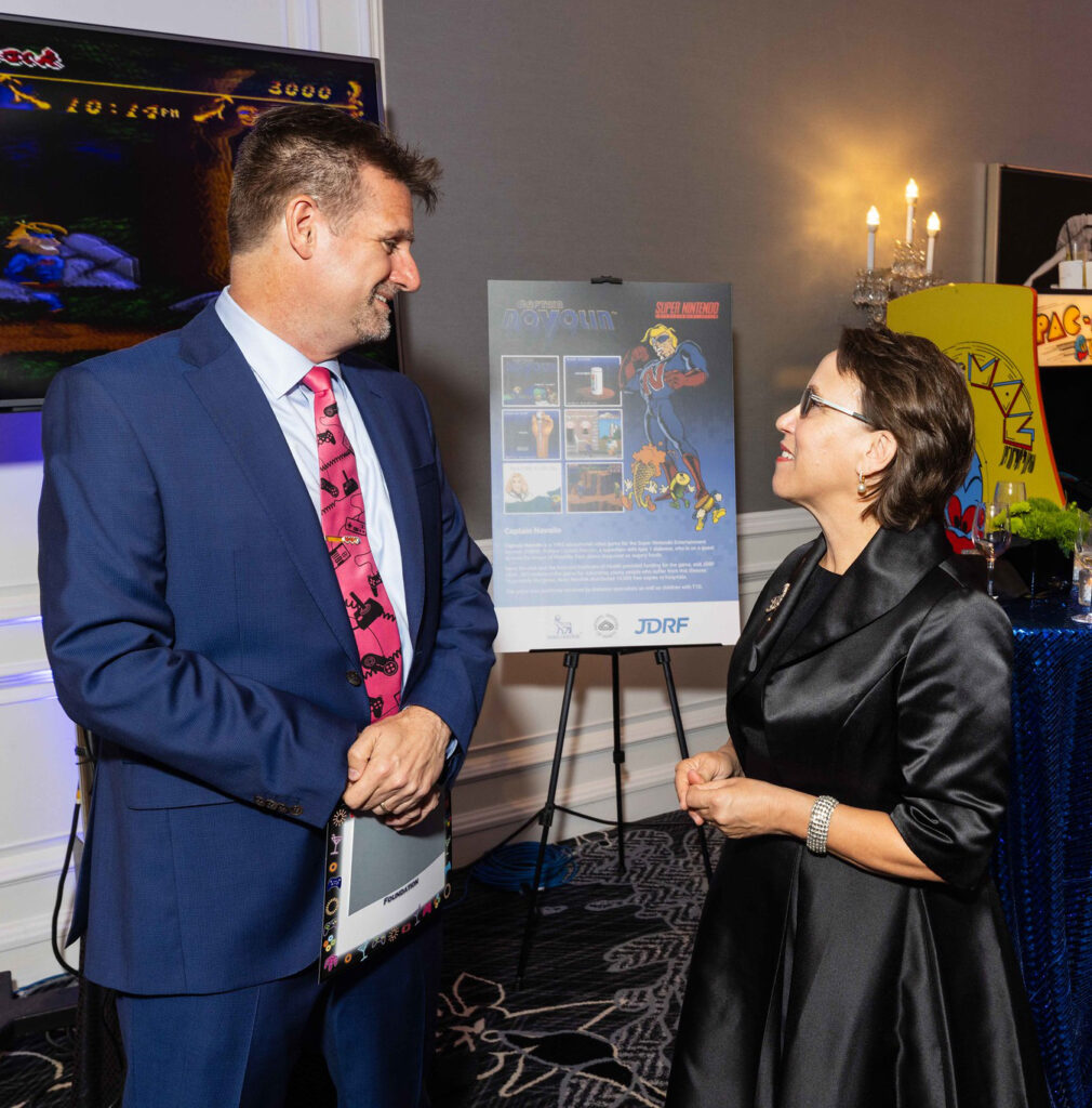 A man in a suit and a woman in a black dress, standing in front of a Captain Novolin poster.