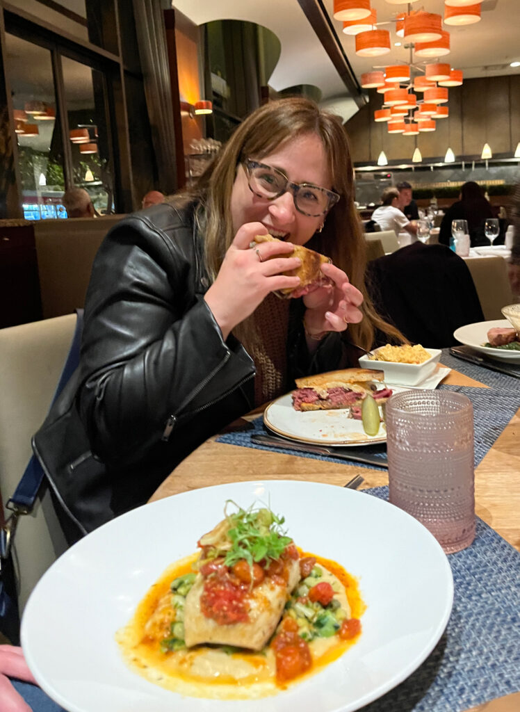 Monique chowing down on a Reuben sandwich, with the restaurant in the background and a salmon plate in the foreground.