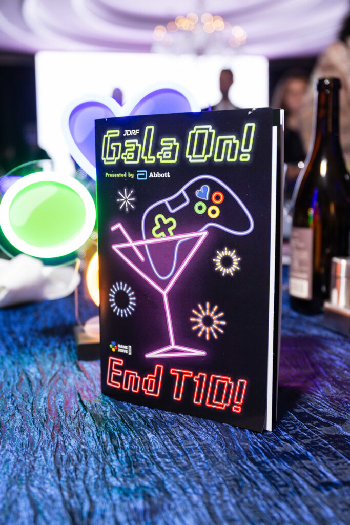 A program standing up on a table with the ballroom visible in the background. The program says "Gala On! End T1D!" and has art of a neon cocktail glass with a game controller in it.