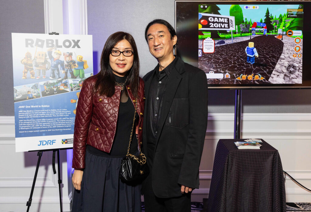 A couple in formal clothing standing in front of a Roblox poster and TV showing Breakthrough T1D One World.